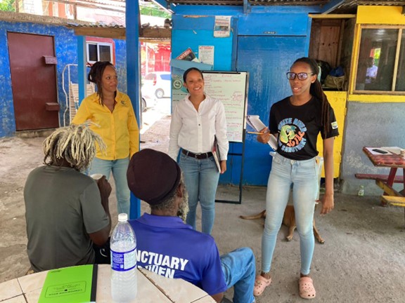 Two women stand and speak to a small group of seated men in an informal outdoor space with bright blue and yellow painted walls.